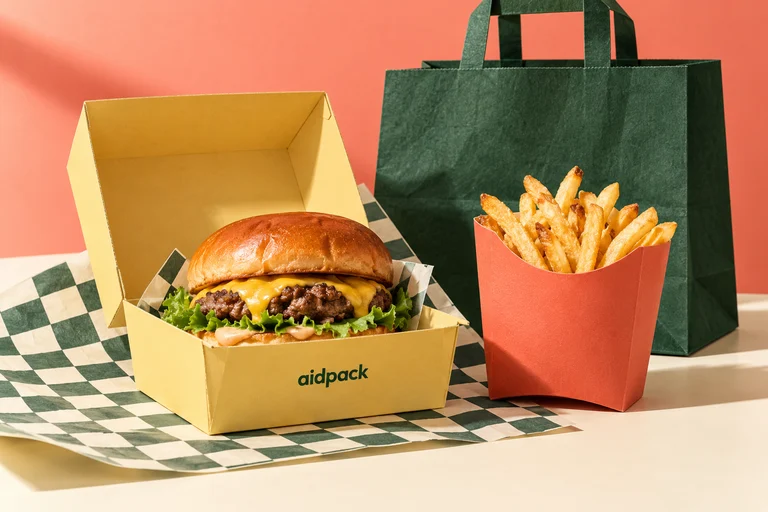 Burger in einer gelben Box, Pommes in einer korallfarbenen Snackbox, bedrucktes Papier und eine grüne Tragetasche.